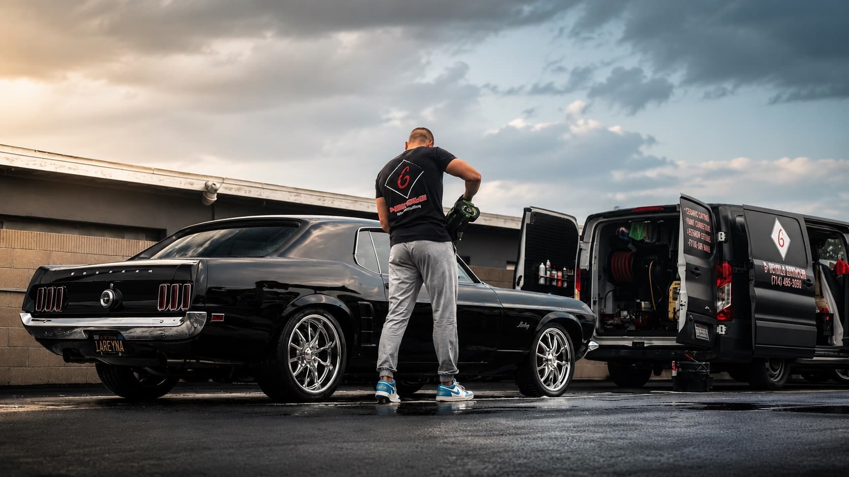 Detailer in a G Mobile Detailing shirt working on a black classic Ford Mustang fastback at sunset with the mobile detailing van in Orange County